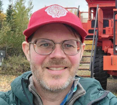 A man with glasses and a red cap smiles for a selfie in front of a large red mining dump truck, with trees and a cloudy sky in the background. He is wearing a green jacket and stands on a grassy and leafy ground.