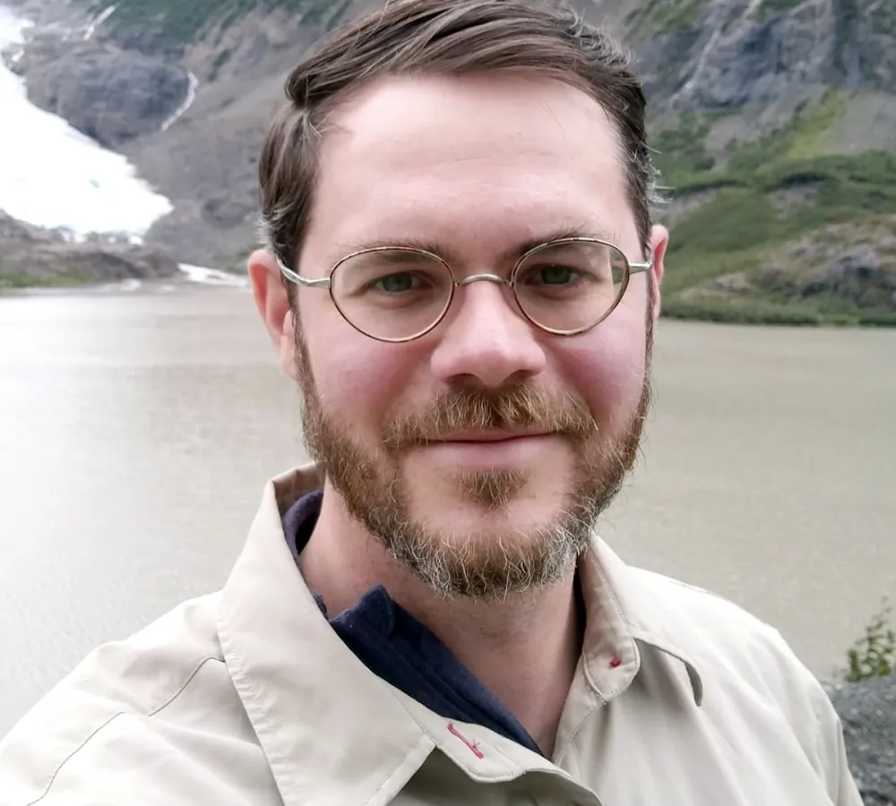 Dr. Patrick Littlejohn wearing glasses, a short beard, and short brown hair smiles at the camera. He is wearing a beige jacket and stands outdoors with a body of water, green hills, and a rocky, snowy mountainside in the background.