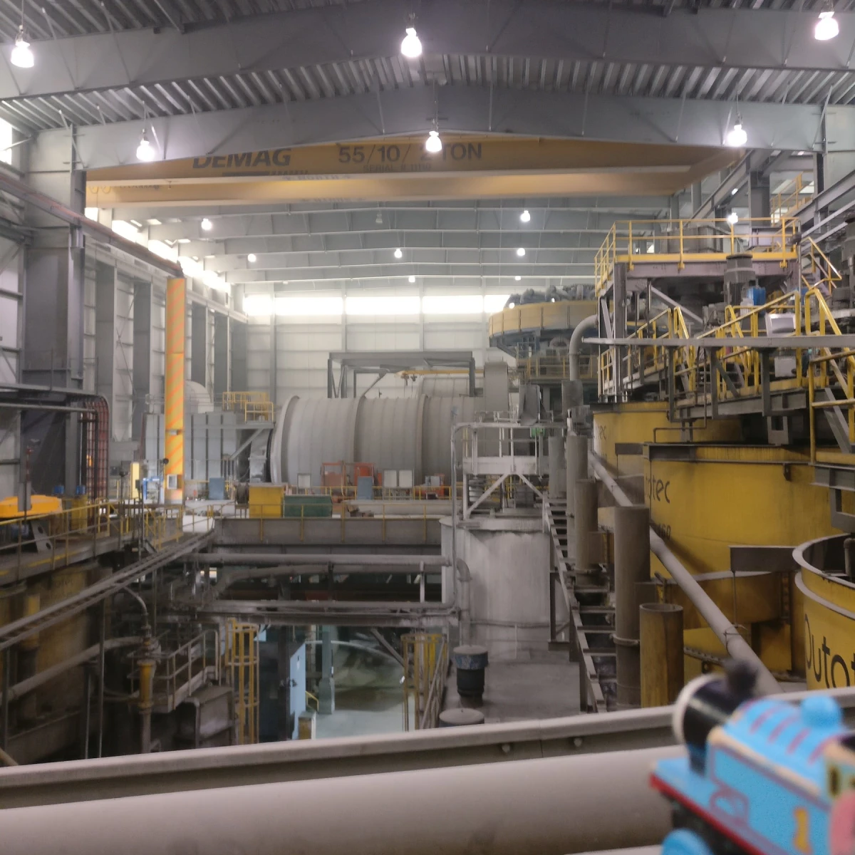 A large industrial factory interior with various heavy machinery, pipes, and metal structures. Overhead lighting illuminates the spacious area. Yellow railings and safety barriers are visible throughout the facility. The environment appears clean and organized.