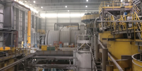 A large industrial factory interior with various heavy machinery, pipes, and metal structures. Overhead lighting illuminates the spacious area. Yellow railings and safety barriers are visible throughout the facility. The environment appears clean and organized.