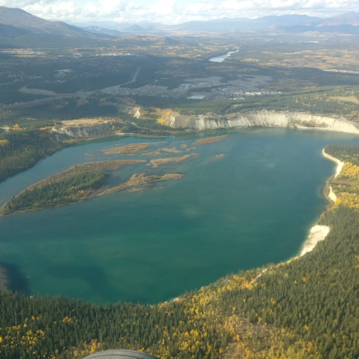 Aerial view of a lake surrounded by land with rugged terrain and forested areas. The water reflects the sky, and the shoreline has sandy beaches. In the distance, there are mountains under a partly cloudy sky. Farmland and scattered buildings are also visible.