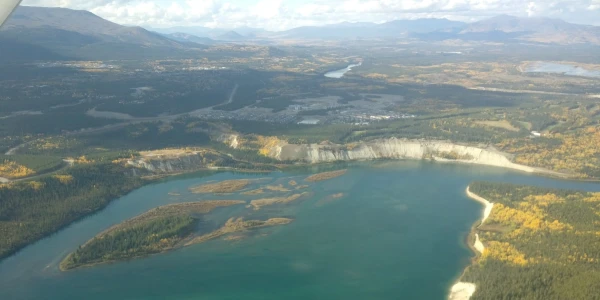 Aerial view of a lake surrounded by land with rugged terrain and forested areas. The water reflects the sky, and the shoreline has sandy beaches. In the distance, there are mountains under a partly cloudy sky. Farmland and scattered buildings are also visible.