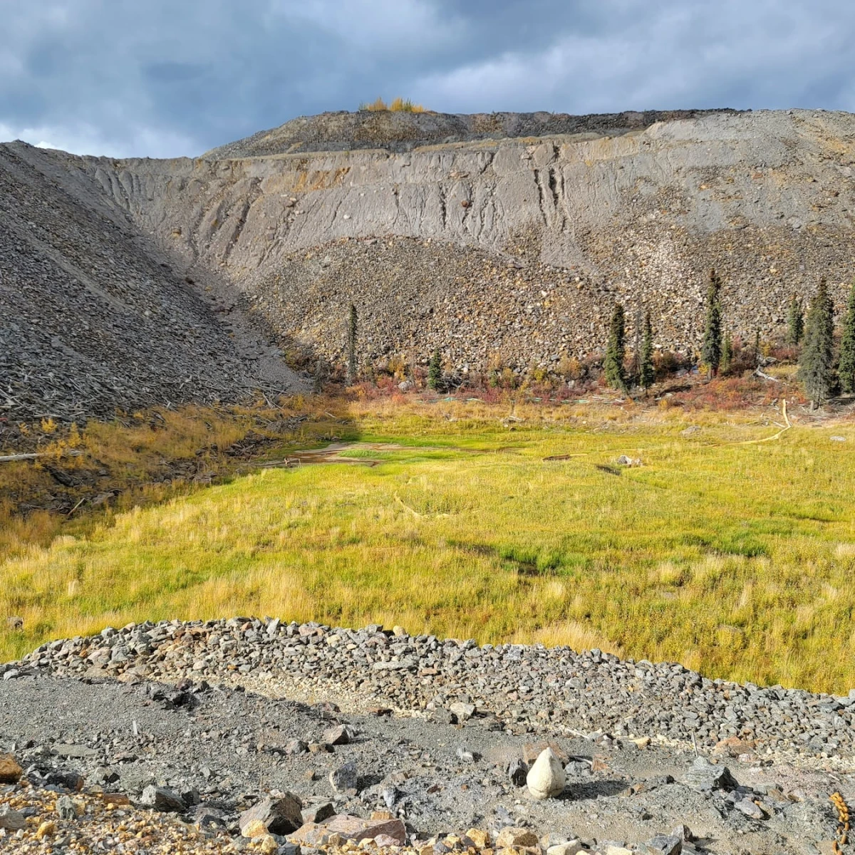 A dry, rocky valley with a patch of green, grassy vegetation in the centre. Sparse trees and shrubs dot the landscape, and the hillsides are covered in loose rocks and gravel. The terrain appears rugged and desolate, with a mix of muted earth tones.