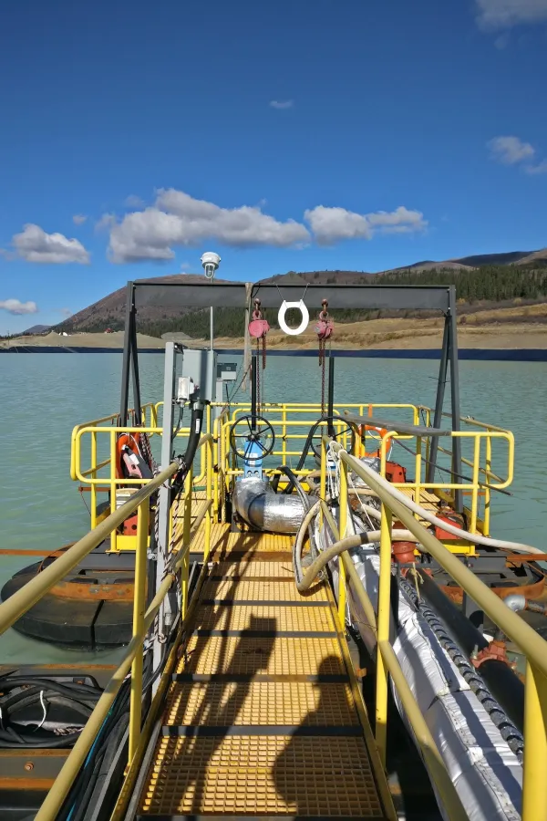 An industrial structure with yellow railings extends over greenish water under a blue sky. The setup includes various cables, hoses, and equipment, set against a backdrop of mountains and forested hills. Shadows of railings and equipment are cast on the walkway.