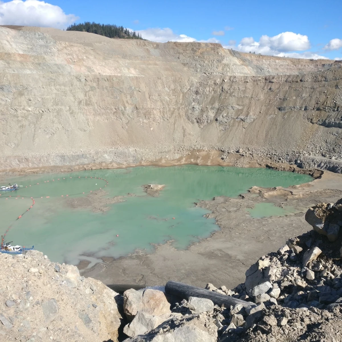 A large, shallow turquoise water pond is visible in a rocky, barren quarry. The steep, jagged walls of the quarry surround the water, and small boats or flotation devices can be seen on the water’s surface near the left side.