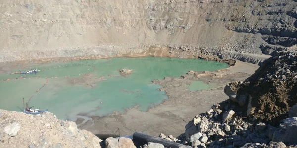 A large, shallow turquoise water pond is visible in a rocky, barren quarry. The steep, jagged walls of the quarry surround the water, and small boats or flotation devices can be seen on the water’s surface near the left side.