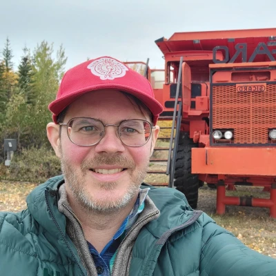 A man with glasses and a red cap smiles for a selfie in front of a large red mining dump truck, with trees and a cloudy sky in the background. He is wearing a green jacket and stands on a grassy and leafy ground.