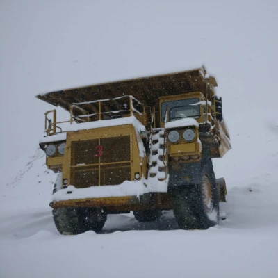 A large, yellow dump truck is covered in snow and parked on a snow-covered surface. The truck has massive tires, and both the vehicle and the surrounding area are dusted with fresh snowfall. The sky appears overcast, suggesting ongoing snow conditions.