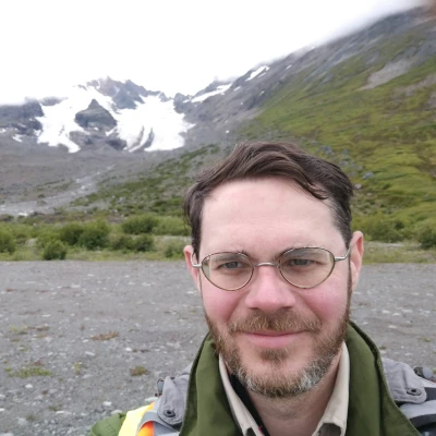 A man with glasses and a light beard takes a selfie outdoors. He is wearing a green jacket with an orange strap over his shoulder. Behind him are snowy mountains with patches of greenery. The sky is overcast.